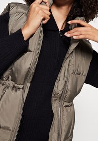 Khaki puffer vest z błyszczącą, gładką teksturą, z zamkiem błyskawicznym i elastycznym pasem, noszony na czarnym swetrze.