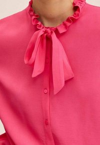 Blusa fucsia brillante con colletto arricciato e un dettaglio con nastro legato al collo. Presenta una chiusura frontale con bottoni e una texture di tessuto morbida.