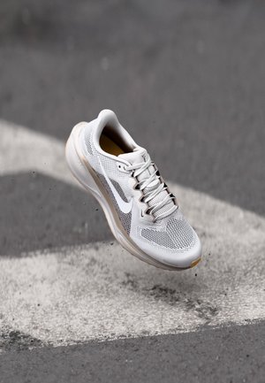 Zapatilla de correr Nike blanca con parte superior de malla y cordones, suspendida sobre asfalto con líneas blancas pintadas y pequeñas piedras dispersas.