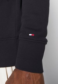 Schwarzer Sweatshirtbund mit geripptem Textil, auf dem ein kleines, gesticktes Logo in Rot, Weiß und Blau zu sehen ist. Sichtbare Nähdetails.