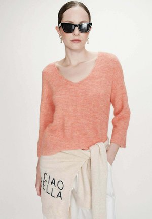 Femme portant des lunettes de soleil noires, des boucles d’oreilles dorées en forme de goutte, un pull corail à col en V, avec un pull crème noué autour de la taille avec l’inscription « CIAO BELLA ».