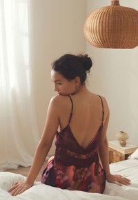 Bordinės gėlėtos naktinės su nėrinių apdaila, permatoma medžiaga, V formos nugara ir reguliuojamomis petnešėlėmis, dėvimos sėdinčio modelio ant lovos.