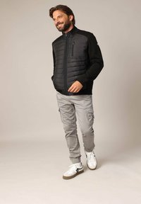 Zwarte pufferjack met een gladde textuur, ritssluiting, lange mouwen en grijze cargobroek. Witte sneakers met zwarte accenten maken de outfit compleet.