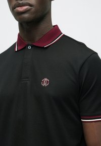 Svart polo-skjorte med en burgunder krage med hvite og røde striper, knapper foran, og en brodert logo på brystet. Strukturert stoff.