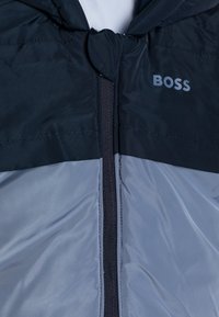 Casaco isolante azul-marinho e cinza com fecho de correr, apresentando um capuz e o logótipo "BOSS" em cinza no lado superior esquerdo. Textura suave.