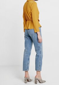 Donna che indossa un top a maniche lunghe di colore giallo senape, jeans chiari a vita alta e scarpe con tacco a blocco a motivo serpente, in piedi di lato.