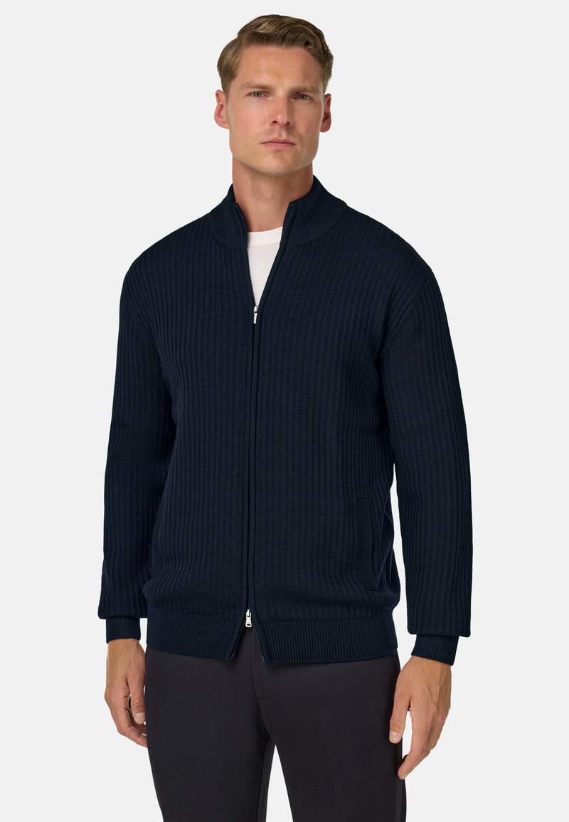 Mornarsko modra pletena zadrga jopica z teksturiranim vzorcem, visokim ovratnikom in stranskimi žepi. Ima dolge rokave ter rebraste manšete in spodnji del.