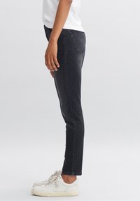 Personne portant un jean slim noir, des baskets blanches et un t-shirt blanc, debout de profil sur un fond blanc uni.