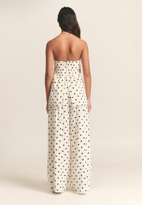 Weißes trägerloses Oberteil mit gerafftem Rücken, kombiniert mit weit geschnittenen Hosen, die ein schwarzes Tupfenmuster aufweisen und eine fließende Silhouette präsentieren.