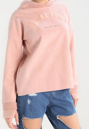 Henkilö, jolla on vaaleanpunainen huppari teksteillä ja revityt siniset farkkushortsit, seisoo yksinkertaisen vaalean taustan edessä.