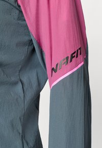 Veste de sport légère, grise et rose, avec une texture finie, arborant un logo "NAFIT" audacieux en noir et des accents roses contrastés.