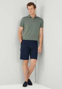 Uomo che indossa una polo verde a maniche corte e pantaloni cargo blu navy, in piedi a piedi nudi contro un muro bianco all'angolo.