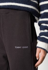 Zbliżenie czarnych spodni z napisem "TOMMY JEANS" obok kieszeni oraz fragment dłoni obok granatowo-białego swetra z dzianiny w paski.