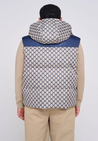 Gilet imbottito con un motivo a rombi in beige e blu. Presenta un cappuccio e spalle blu solide, con una finitura liscia e testurizzata.