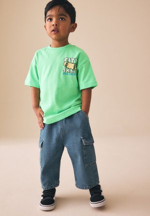 Băiat tânăr în picioare, purtând un tricou verde "City Skate", pantaloni cargo largi din denim și adidași negri cu tălpi albe.