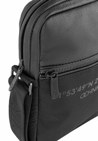 Czarna torba wykonana ze skóry i teksturowanej tkaniny, z podwójnymi zamkami błyskawicznymi, wyraźnym logo i nadrukowanymi współrzędnymi po boku.