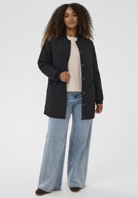 Frau mit lockigem Haar, die eine schwarze gesteppte Jacke, einen beigen Pullover, hellblaue Jeans mit weitem Bein und braune Schuhe trägt, steht vor einem schlichten Hintergrund.