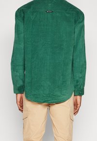 Zelená manšestrová košile s dlouhými rukávy, manžetami na knoflíky a rovný lem; má texturovaný povrch a značkovou cedulku u výstřihu.