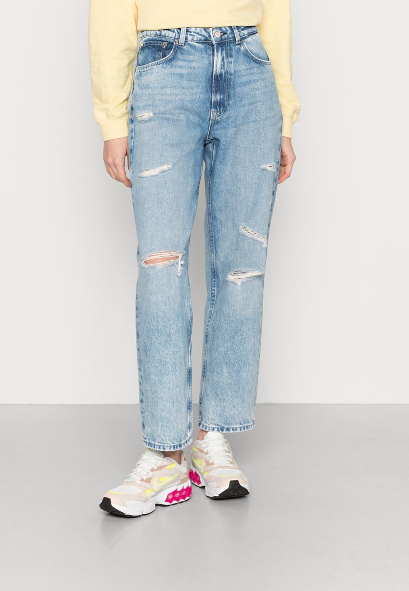 Personne portant un jean déchiré bleu clair et des baskets blanches avec des touches roses et jaunes, debout sur un sol gris.