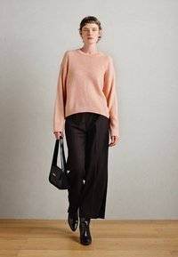 Pull en tricot rose clair avec une coupe décontractée, associé à un pantalon noir à jambes larges. Le modèle tient un sac à main noir ; elle porte des chaussures noires à bout pointu.