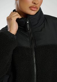 Schwarze Zip-Jacke mit einer Mischung aus glatten und strukturierten Materialien. Mit hohem Kragen, Reißverschluss und weichen Fleece-Details an den Ärmeln.