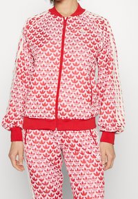 Röd mönstrad jacka med dragkedja framtill, elastiska muddar och fåll. Har ett repetitivt lövmönster i nyanser av rött och rosa.