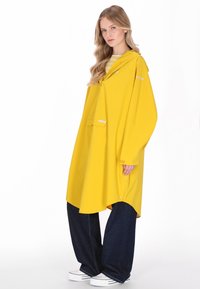 Pelerină de ploaie galbenă cu glugă, croială supradimensionată, cu un buzunar frontal și detalii cu nasturi. Poartă peste pantaloni largi de culoare albastru închis.