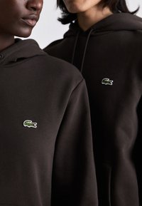 Sudaderas marrones con una textura suave, que cuentan con un pequeño logo bordado de un cocodrilo verde y blanco en el pecho.