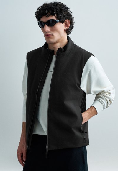Gilet nero senza maniche con colletto alto e zip; indossato sopra una felpa grigio chiaro con maniche a contrasto; texture liscia.