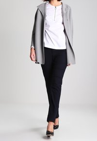 Cappotto grigio in misto lana con ampio colletto e chiusura a un bottone, indossato sopra una camicia bianca e pantaloni neri, completato da ballerine nere.