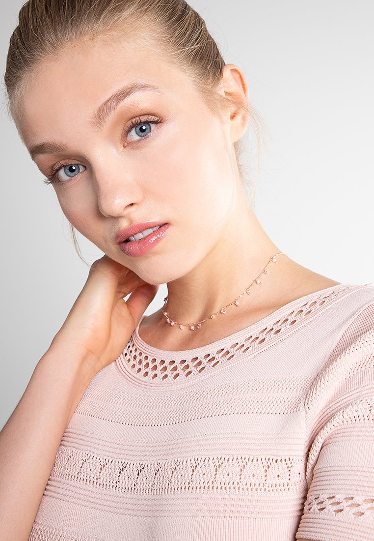 Zarte Choker-Halskette mit kleinen, klaren Edelsteinen, die auf einem hellrosafarbenen, strukturierten Oberteil liegt, das offene Strickdetails und horizontale Streifen aufweist.