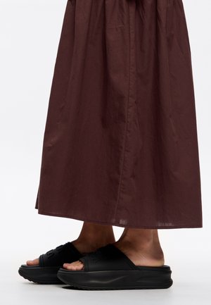 Osoba nosząca długą brązową spódnicę i czarne klapki na platformie, stojąca na białej powierzchni.