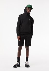 Jeune homme portant un sweat à capuche noir, un short noir, des chaussettes noires, des baskets noires et blanches et une casquette de baseball verte, souriant et debout sur fond blanc.