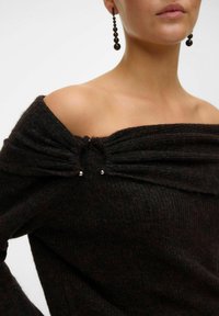 Svart off-shoulder stickad tröja med samlad tyg vid halsringningen och två metalliska detaljer. Mjuk textur med en något lös passform.