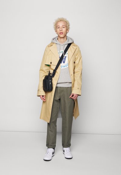 Beige trenchcoat over en grå hoodie med logo, grønne bukser og hvide sneakers med sorte accenter. Sort crossover-taske på skulderen.