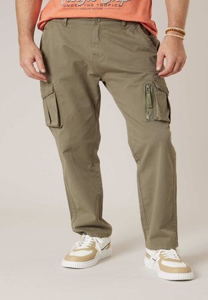 Homme portant un pantalon cargo vert olive avec plusieurs poches et des baskets beige-blanc, debout devant un fond neutre.