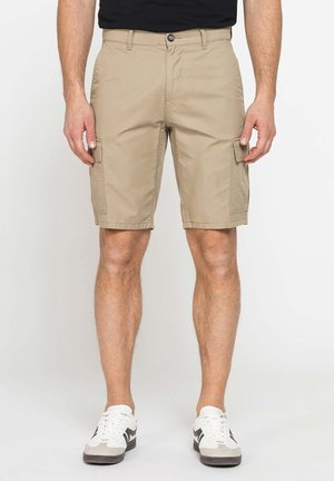 Uomo con pantaloncini cargo beige con tasche frontali e scarpe da ginnastica bianche, in piedi davanti a uno sfondo uniforme.