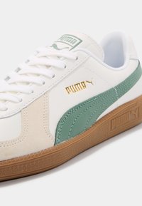 Basket blanche Puma avec bande latérale verte, accents en daim beige, logo Puma doré, lacets blancs et semelle en gomme.