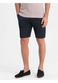 Tummansinisiä shortseja, jotka on valmistettu tekstuurista kankaasta, sivutaskuilla ja keskipituisina reisiin saakka, yhdistettyinä mustiin samettisiin loafer-kengiin.