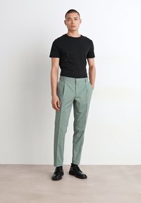 T-shirt noir ajusté associé à un pantalon taillé vert clair. Le pantalon présente des poches avant et une texture lisse ; porté avec des chaussures noires.