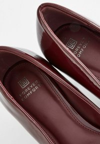Küpsetatud lakknahast burgundia värvi kõrged kontsad, siledat pinnatöötlust, ümarad varbad, pehmendava sisetallaga ja hõbedane "Forever Comfort" logo interjööris.