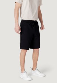 Black cotton shorts with an elastic waistband and drawstring, featuring side pockets and a knee-length cut. Paired with white sneakers.