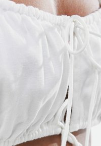 Vit avklippt topp gjord av texturerad tyg. Har elastiskt veck vid toppen och justerbara band på sidorna. Slät och lättviktig.