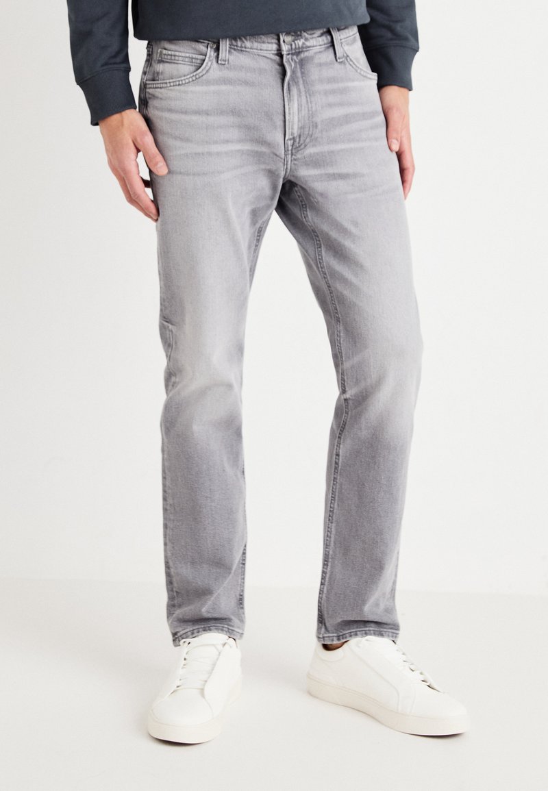 Vaqueros de mezclilla gris claro con corte recto, cinco bolsillos y una cintura clásica, combinados con zapatillas blancas. Textura suave y mínimo desgastado.