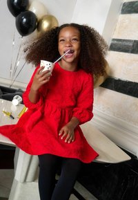 Sarkans, teksturēts kleita ar puffētiem piedurknēm, apvienota ar melniem zeķubiksēm. Bērns tur polka punktiņu kausu ar salmiņu, smaidot, uz marmora galda.