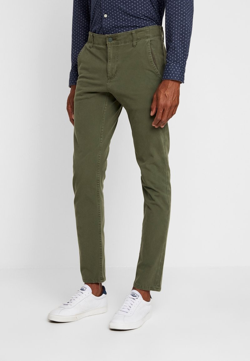 Homme portant un pantalon slim kaki, une chemise boutonnée bleu marine à motifs, et des baskets blanches, debout devant un fond uni.