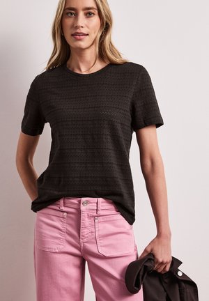 Kvinde iført en sort tekstureret kortærmet top og lyserøde bukser, der holder en sort jakke i højre hånd mod en ensfarvet baggrund.