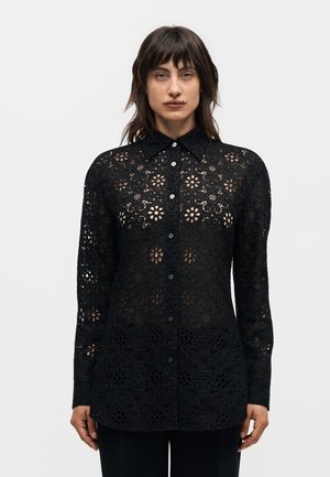 Femme portant une chemise noire à manches longues en dentelle florale avec boutons, debout devant un fond clair uni.