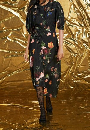 Robe noire à motifs floraux avec manches bouffantes, taille froncée et motifs floraux vibrants. Portée avec des collants en dentelle noirs et des chaussures à talons. Fond doré.