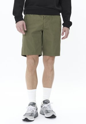 Olivgrüne Shorts aus strukturiertem Stoff, mit geradem Schnitt, Seitenstaschen und lässiger Passform, kombiniert mit grauen Turnschuhen und weißen Socken.